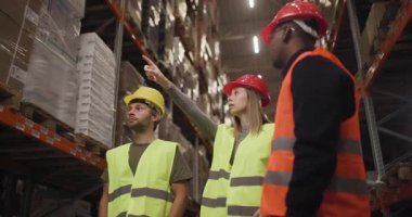 Yansıtıcı ekipmanlı depo çalışanları uzun raflardaki stokları inceliyor. Lojistik bir ortamda envanter yönetimi ve güvenlik uygulamalarına odaklanan takım işbirliği.