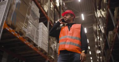 Portakal rengi güvenlik yeleği giyen depo çalışanı ve rafların arasında telefonla konuşan, endüstriyel bir depolama ortamında lojistik, iletişim ve takım çalışmasını ön plana çıkaran bir şapka..