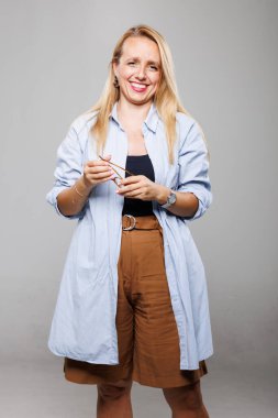 Açık mavi gömlek ve kahverengi şort giyen bir iş kadını, elinde bir çift gözlük tutarak güvenle ayakta duruyor. Bir stüdyo ortamında dostane bir tavır sergiliyor..