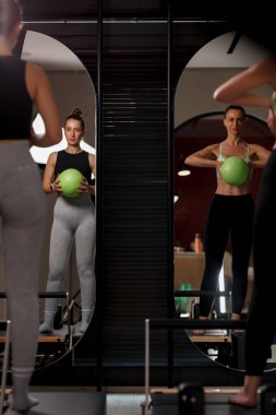 Doğal ışıkla dolu modern bir spor salonunda iki kadın, yol gösterici olarak aynaları kullanırken kendi formlarına odaklanmış bir şekilde yeşil bir spor topuyla güç antrenmanı yapıyorlar..