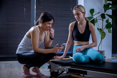 Modern bir spor salonunda iki kadın telefonlarını kontrol ederken aynı anda gülüyorlar. Antrenman kıyafeti giymişler ve etrafları pilates yenilikçiliği ekipmanlarıyla çevrili..