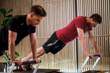 Çağdaş bir fitness stüdyosunda iki birey, antrenman sırasında güç, denge ve esnekliği vurgulayarak, reformcu pilates makineleri üzerinde gelişmiş hareketler gerçekleştiriyor..