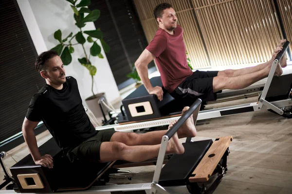 İki adam, doğal dekoru ve yeşilliği olan iyi ışıklandırılmış bir fitness stüdyosunda güç ve esneklik üzerine çalışan reformcu bir pilates antrenmanında..