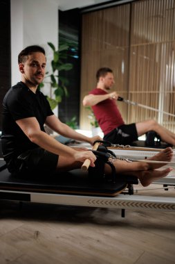 İki kişi şık bir stüdyoda pilates antrenmanı yapıyor, akşam saatlerinde güçlerini ve esnekliklerini arttırmak için yenilikçi makineler kullanıyorlar..