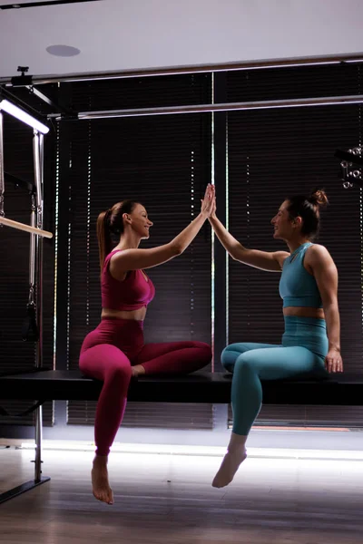 İki kadın spor salonundaki bir bankta yan yana oturur, ellerini büyük pencerelerden gelen doğal ışıkla çevrili, eğlenceli bir fitness egzersizinde birleştirirler..