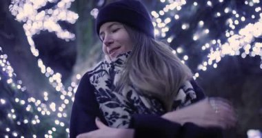 Sıcak bir eşarp ve şapka takmış bir kadın soğuk bir kış akşamında güzel aydınlatılmış bir ağacın yanında gülümser. Şehrin neşeli ve neşeli havasını yakalar..