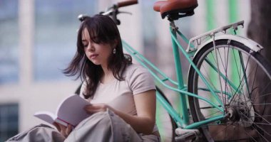 Kitabına kendini kaptırmış genç bir kadını kentsel bir zemine karşı şık turkuaz bisikletinin yanında otururken gösteren sakin bir görüntü..