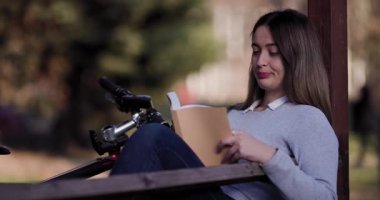 Güneşli bir parkta bisikletinin yanında oturmuş kitap okumaya dalmış genç bir kadının huzurlu bir sahnesi..