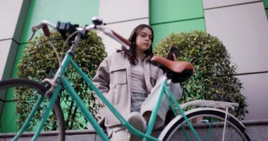 Akıllı telefonunu kullanırken gülümseyen sıradan bir genç kadın, klasik bisikletinin yanında modern bir şehir arka planına karşı oturuyordu..