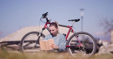 Odaklanmış genç bir kadın çimlerin üzerinde kitap okumaktan hoşlanır, bisikleti yakınlarda, açık mavi bir gökyüzünün altında park halindedir..