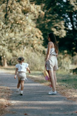 Bir kadın ve genç kızı yemyeşil bir parkta gölgeli bir yolda birlikte güneşli bir günün tadını çıkarıyorlar..