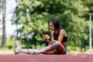 Pistte oturan genç bir kadın, akıllı telefonuna odaklanmış, güneşli bir günde güneşli bir spor salonunda mola veriyor..