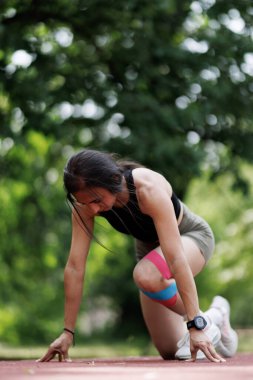 Güneşli bir pistte koşuya hazırlanan atletik kıyafetli bir bayan sporcu. Odaklanmış ifadesi ve dengeli duruşu kararlılık ve uygunluğu vurguluyor..