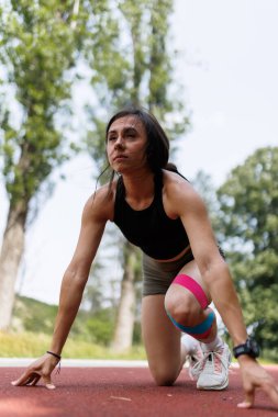 Kararlı bir atlet, yeşil, doğal bir çevre ile çevrili, hızlı koşmaya hazır, açık hava pistinin başlangıç çizgisinde çömelir..