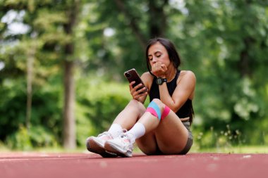 Koşu pistinde oturan genç bir kadın akıllı telefonuna bakıyor. Atletik kıyafetler giyiyor. Bacağında da renkli bir bant var. Açık havada sakin bir ortamda..