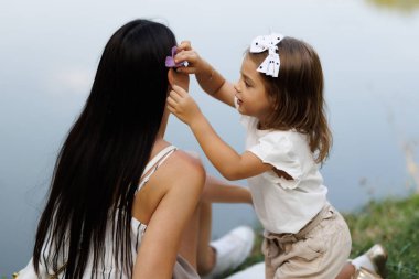 Güneşli bir günde güneşli bir gölün kenarında otururken annesinin saçına toka takan küçük bir çocuğu gösteren dokunaklı bir sahne aile bağlarını ve sevgisini gösteriyor..