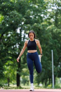 Spor elbiseli bir kadın, yeşil ağaçlar ve güneş ışığıyla çevrili, açık havada koşan bir pistte esneme egzersizleri yapıyor. Spor, sağlık ve açık hava aktiviteleri olay yerini somutlaştırıyor..