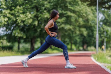 Yeşil ağaçlarla çevrili bir pistte esneme egzersizi yapan bir kadın. Açık bir günde spor, sağlık ve etkin bir yaşam tarzını teşvik ediyor..