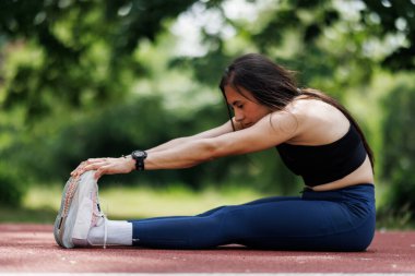 Kendini adamış bir bayan sporcu, güneşli bir günde çevresi yeşilliklerle çevrili açık havada konsantrasyonuyla geriniyor. Formun, sağlığın ve sağlıklı bir yaşam tarzının mükemmel bir tasviri..