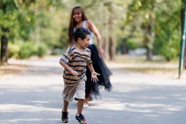 Genç bir çocuk, neşeli bir şekilde parkta koşarken, gülümseyen bir kadın onu hevesle takip ediyor, açık havada neşeli ve neşeli bir sahne yaratıyor..