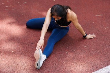 Atletik giyinmiş bir kadın, kırmızı bir stadyum pistinde otururken bacağını uzatarak, esnekliği ve egzersiz ve sporla ilgili aktiviteleri geliştirmeye odaklanıyor..