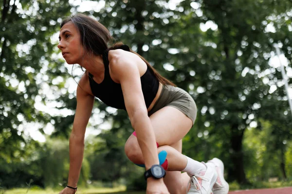 Parkta çömelmiş spor yapan genç bir kadın, spor kıyafetleri, akıllı bir sepet, ve diz desteği, gücü, dayanıklılığı ve aktif yaşamı sembolize ediyor..