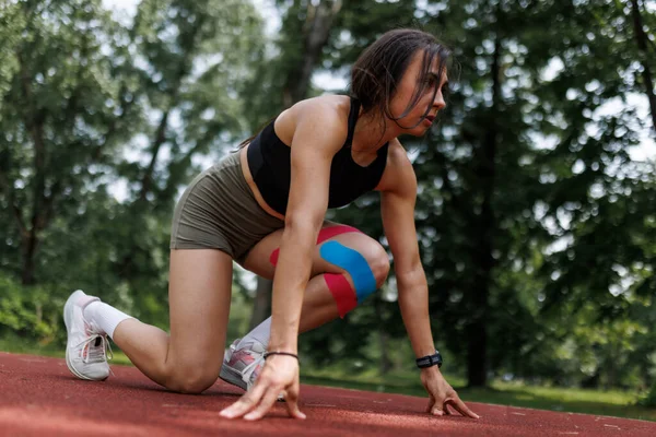 Yeşilliğin ortasında kırmızı bir pistte koşmaya hazır kararlı bir bayan atlet. Dizine kineziyoloji bandı takıyor. Bu da fiziksel aktivite sırasında yaralanma desteğini gösteriyor..