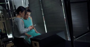 İki birey, loş bir stüdyoda antrenman yapmaya odaklanmış durumda. Biri antreman aletinin üzerine oturmuş telefona bakarken, diğeri de yakınlarda durup gözetliyor..