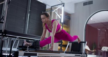 Modern bir fitness stüdyosunda, bir birey etkileyici bir esneme hareketi yapar ve bir pilates reformcusu üzerinde egzersiz yapar. Oturumda şık bir ortamda güç ve esneklik eğitimi veriliyor.