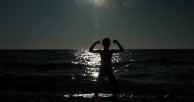 A striking silhouette of a young child celebrating freedom and hope on a tranquil beach as the sun sets over the glistening ocean waves, hands raised in triumph.