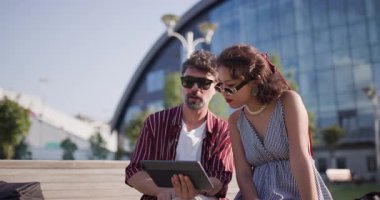 Bir kadın ve erkek, şehir merkezindeki bir bankta otururken tabletle mutlu bir şekilde etkileşime geçiyorlar. Rahatlar ve gülümserler..