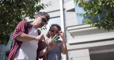 Açık havada akıllı telefon kullanan bir adam ve kadın güneşli bir günde sohbet ediyorlar. Bir binanın yanında duruyorlar, teknolojiyle etkileşime geçiyorlar..