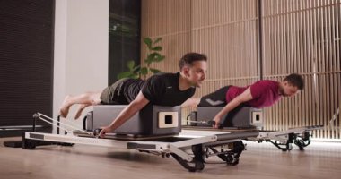 Gösterişli, iyi ışıklandırılmış bir stüdyoda, iki kişi yoğun bir pilates yenilikçisi eğitimi alıyor, güç ve esnekliğe odaklanıyor. Atmosfer enerjik ve motive edici..