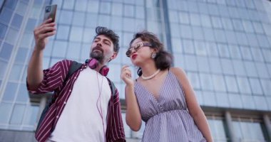 Çağdaş bir cam binanın dışında selfie çeken neşeli genç bir çift mutluluğu ve modern yaşam tarzını gözler önüne seriyor..