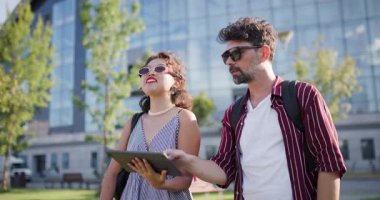 Güneşli bir günde dışarıda bir tablet paylaşan mutlu çift, birlikte boş vakitlerin tadını çıkarırken çağdaş bir binanın yanında duruyorlar..