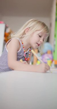 Genç bir kız odasındaki masada pastel kalemlerle mutlu bir şekilde resim çizer. Renkli kolyesi neşe dolu atmosfere katıyor..