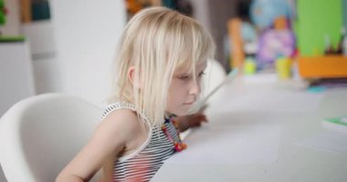 Genç bir kız beyaz bir masada oturuyor, kalemle kağıt parçaları çiziyor. Sanat malzemeleri ve bir küre de dahil olmak üzere çeşitli rengarenk eşyalar, etrafını sarın..