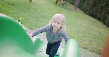 Küçük sarışın bir kız oyun parkında yeşil bir kayağa tırmanır. Eğleniyor ve gülümsüyor..