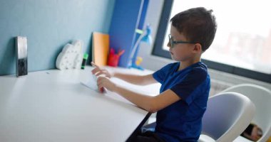 Genç bir çocuk evdeki masasında oturuyor ve dikkatlice origami kağıdını katlıyor. Odaklanmış durumda ve zanaat faaliyetlerini seviyor..