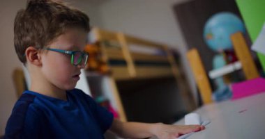 Gözlüklü genç bir çocuk yatak odasında masasında oturur ve bir kağıt uçak katlar. Uçuruyormuş gibi yapmadan önce havada tutuyor ve inceliyor..