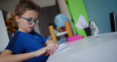 Gözlüklü genç bir çocuk masasına oturur ve dikkatlice bir kağıt parçası katlar, muhtemelen bir origami oluşturur..