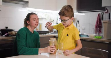 Genç bir çocuk annesi izlerken bir bardak suya dikkatlice çikolata tozu ekler. Mutfakta çikolatalı süt yapıyorlar..