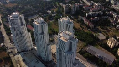 Kentsel bir bölgedeki üç modern gökdelenin havadan görünüşü. Çevresindeki manzarada alçak binalar, yeşil alanlar ve yollar yer alıyor..