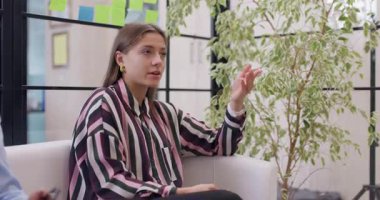 Modern bir ofiste beyaz bir koltukta oturan genç bir kadın ekranın dışında biriyle konuşurken el kol hareketi yapıyor..