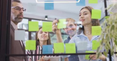 İş arkadaşları başlangıç projesi fikirlerini tartışıyor ve beşlik çakıyor. Cam duvarda yapışkan notlarla beyaz iş adamları. Renkli çıkartmalı Scrum. Düşük açı görünümü.