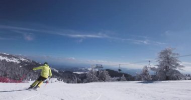 Bir kayakçı, uzun ağaçlar ve engin mavi gökyüzüyle çevrili beyaz karla kaplı bir dağ yamacından ustalıkla iner. Parlak kış güneşi kayak yapmak için mükemmel manzarayı güzelleştirir..