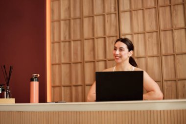 Dost canlısı bir kadın şık bir resepsiyonda oturmuş, çağdaş bir ofis ortamında öğleden sonra lambasının keyfini çıkarırken bilgisayarını meşgul ediyor. Hoş bir gülümsemesi var..