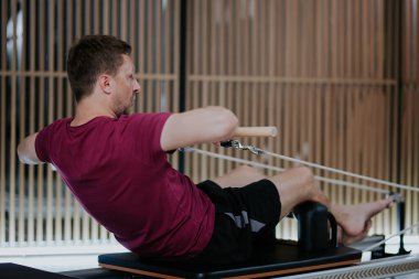 Bir adam, zarif ve iyi ışıklandırılmış bir fitness stüdyosunda, çekirdek kuvvete ve esnekliğe odaklanarak, reformcu bir pilates egzersizi yapıyor. Çevre sağlıklı bir çalışma atmosferini teşvik eder..