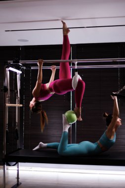 Çağdaş bir stüdyoda, iki kadın pilates egzersizi yapıyor. Biri teçhizatın üzerinde baş aşağı asılı dururken, diğeri yeşil bir top dengeliyor, güç ve koordinasyon gösteriyor..