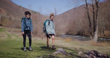 İki bayan arkadaş nehir kenarında yürüyüş yapıyorlar, sırt çantaları taşıyorlar ve macera için giyinmişler..